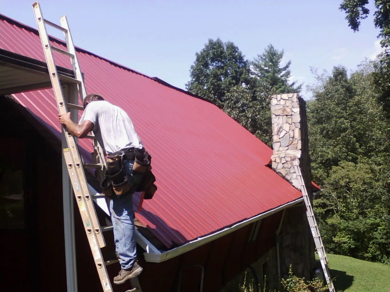Roofer on a ladder beside a red metal roof and stone chimney — Soffit & Fascia Repair by Roofing Brothers in Rolla